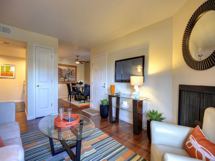 Living Room Facing Entrance with Hardwood Inspired Floor, Rug, Glass Table, Round Wall Mounted Mirror, White/Gray Sofa, and Flat Screen Television